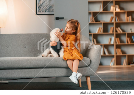 Cheerful little girl is with maltese dog indoors in domestic room on the sofa Cheerful little girl is with maltese dog indoors in domestic room on the sofa 126170829