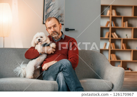Man is sitting with his maltese dog on the sofa indoors 126170967