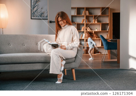 Woman with book, girl is having fun. Mother with her daughter is together in the domestic room Woman with book, girl is having fun. Mother with her daughter is together in the domestic room 126171304