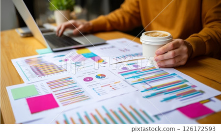 Person working on business reports with laptop and coffee in a bright workspace during the morning 126172593