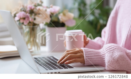Person works on laptop while holding coffee cup in cozy environment with floral arrangement during daytime 126172848