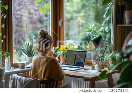 Young woman is working from home, sitting at her desk in front of a laptop, in a bright and cozy home office full of plants 126173891