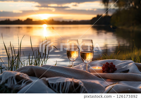 Two glasses of white wine and grapes on a blanket by the lake, creating a romantic atmosphere at sunset 126173892