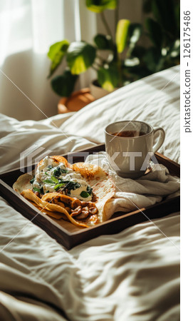 Mini crepes with cashew cream, spinach, and mushrooms sit on a dark wooden tray, next to a steaming cup of mint tea. The scene is set on a bed with soft linens in a modern, bright bedroom 126175486