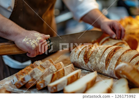 Baker is slicing a loaf of freshly baked bread on a wooden board, with slices of bread scattered around him 126175992