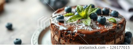 Chocolate cake topped with fresh blueberries and mint leaves on a silver plate in a cozy kitchen setting 126177015
