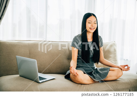 Asian woman finds balance meditating in lotus pose with laptop on sofa. Serene businesswoman practices relaxation and mindfulness while working online. Portrait of a smiling student in harmony. Asian woman finds balance meditating in lotus pose with laptop on sofa. Serene businesswoman practices relaxation and mindfulness while working online. Portrait of a smiling student in harmony. 126177185