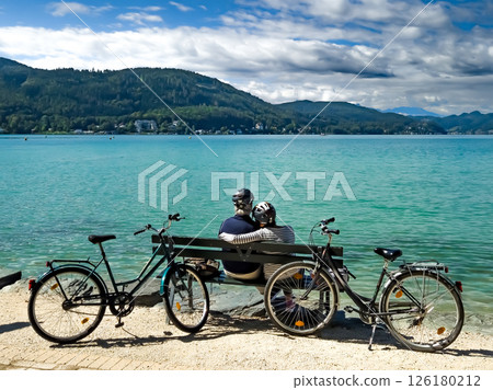 Sporty Senior Couple With Bicycles Sit On Bench At Lake Woerthersee In Carinthia, Austria 126180212