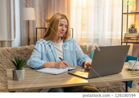 Young woman studying at home video laptop lesson focus, notes in notebook, attentive learning moment 126180594