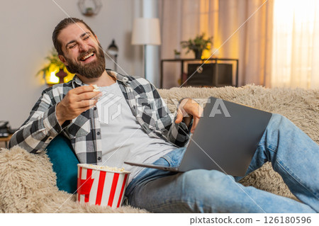 Young man relaxing at home sofa, eating popcorn bowl, laptop laughter, video, comedic fun enjoyment 126180596