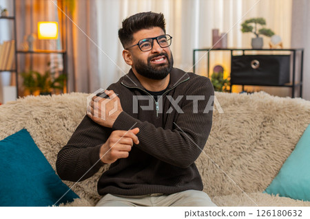 Indian man sitting at home holds shoulder with discomfort, reacting to physical pain and tension 126180632