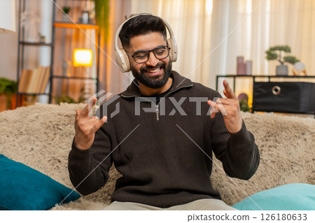 Indian man smiling listens to energetic music in wireless headphones, shows rock-n-roll gesture Indian man smiling listens to energetic music in wireless headphones, shows rock-n-roll gesture 126180633