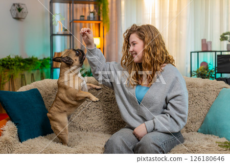 Young woman training pug dog on home sofa, pet standing on hind legs and posing with cute attention 126180646