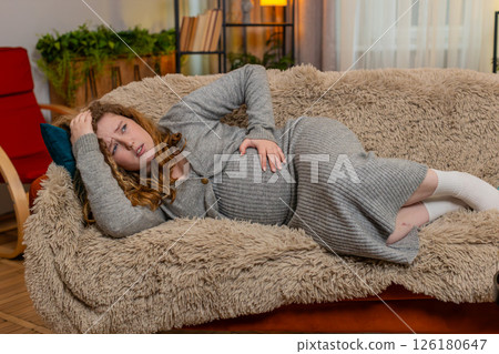 Pregnant young woman lying on home sofa holding belly wincing in pain discomfort worried expression 126180647