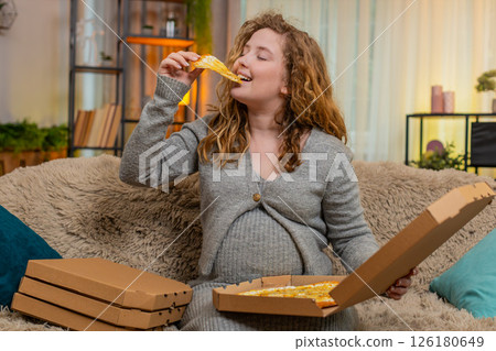 Pregnant woman opening pizza box at home, tasting slice with satisfied smile, enjoying comfort food 126180649