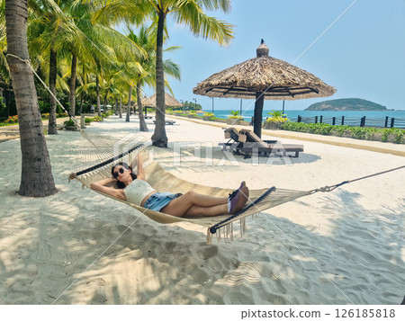 Brunette Russian girl relaxing in a beach hammock wearing denim mini skirt and t-shirt with sunglasses. Nha Trang beach with sunbeds and palm trees, Vietnam 126185818
