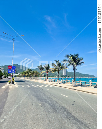 Empty clean road in Nha Trang during siesta time with traffic signs, blue sea featuring traditional Vietnamese boats, palm trees and sunny sky 126185824