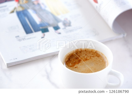 Image of a man reading a magazine while drinking a cafe latte (a mixture of espresso and milk) at a white table 126186790