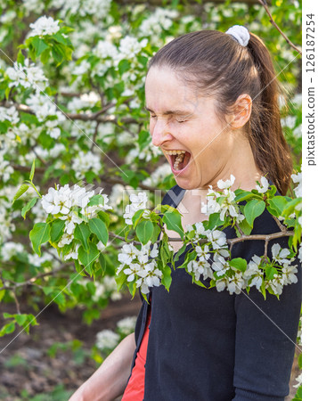 Young woman sneezes in the park against the background of a flowering tree. Allergy to pollen concept. Young woman sneezes in the park against the background of a flowering tree. Allergy to pollen concept. 126187254