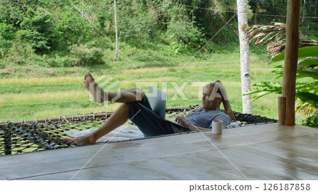 A young man working in a hammock, engaging in remote work on his laptop, surrounded by lush tropical jungle, he enjoys the serenity and beauty of nature on a terrace A young man working in a hammock, engaging in remote work on his laptop, surrounded by lush tropical jungle, he enjoys the serenity and beauty of nature on a terrace 126187858
