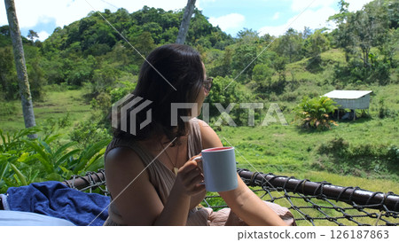 Young woman relaxes in a hammock on a tropical terrace, savoring freshly brewed coffee while taking in breathtaking views of rice fields and lush jungle greenery Young woman relaxes in a hammock on a tropical terrace, savoring freshly brewed coffee while taking in breathtaking views of rice fields and lush jungle greenery 126187863