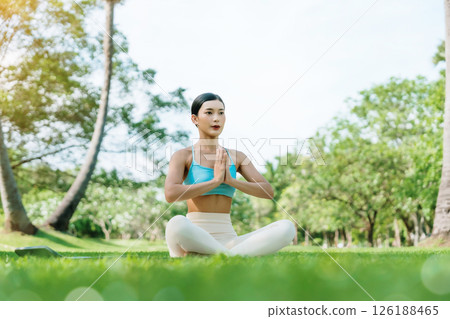 Asian woman meditating in a peaceful park setting, sitting cross-legged on green grass with trees 126188465