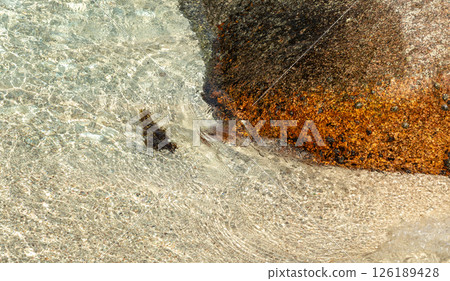 Clear ocean water flows over textured sand and orange granite rock Clear ocean water flows over textured sand and orange granite rock 126189428