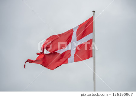 The Denmark flag flutters proudly on a flagpole, displaying its red background and white cross against a cloudy, overcast sky. The atmosphere is calm and reflective. 126190967