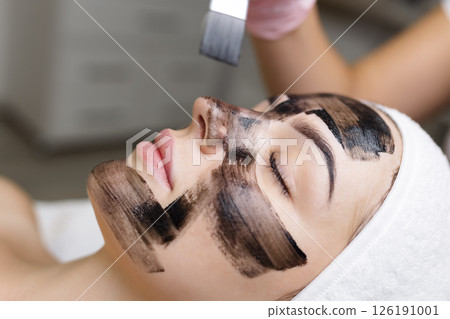 cosmetologist applying black mask on pretty woman face wearing black gloves, gorgeous woman in spa having facial procedures cosmetologist applying black mask on pretty woman face wearing black gloves, gorgeous woman in spa having facial procedures 126191001