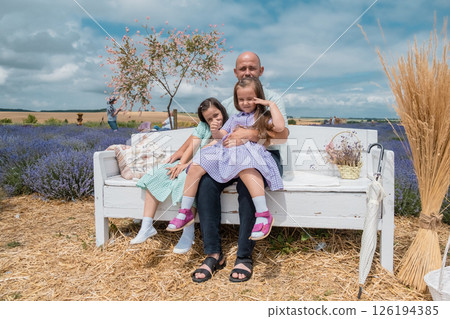 A father and two daughters in beautiful dresses resting on a bench in a lavender field. 126194385