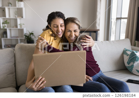 Excited mature mum teen daughter hugging on sofa opening parcel Excited mature mum teen daughter hugging on sofa opening parcel 126198184