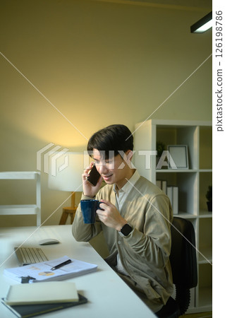 Young entrepreneur holding cup of coffee and talking on mobile phone at home office desk 126198786