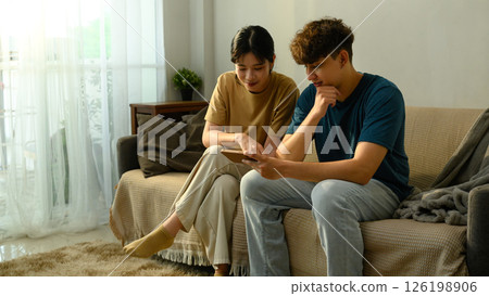 Young couple sitting on sofa and browsing smartphone together in living room Young couple sitting on sofa and browsing smartphone together in living room 126198906