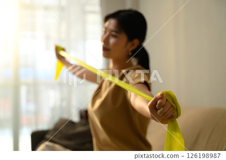 Asian woman using resistance band for arm exercises in a bright living room 126198987