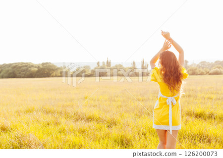 Slender woman in yellow summer dress walking in wheat field at sunset 126200073