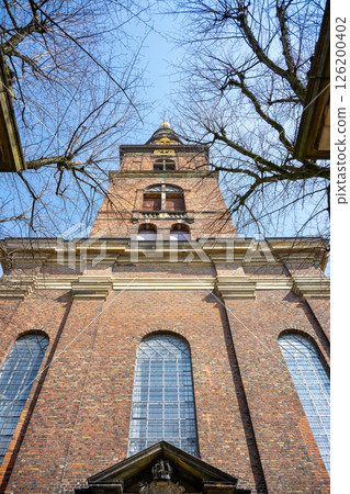 Visitors admire the impressive architecture of the Church of Our Saviour in Copenhagen, Denmark. The towering spire and intricate brickwork captivate. The clear sky enhances the beauty. 126200402