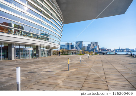 Visitors take advantage of a sunny day at Copenhagen Opera House, strolling along the building's modern architecture and enjoying views of the waterfront and cityscape. 126200409