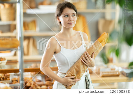 Pretty young girl buyer chooses fresh pastries in bakery, standing with baguette in front of display case with baked goods in bakery market 126200888