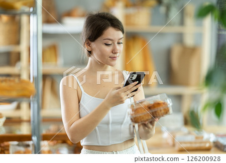 Cute young girl choosing pastries in bakery store, scans QR code on packaging box using mobile Cute young girl choosing pastries in bakery store, scans QR code on packaging box using mobile 126200928