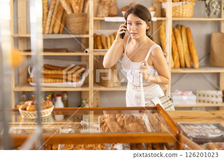 Nice young girl shopper talking on phone while choosing pastries in bakery shop Nice young girl shopper talking on phone while choosing pastries in bakery shop 126201023
