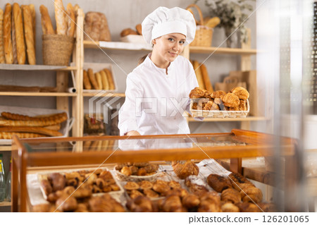 Young woman employee puts croissants in window, arranges display of goods at bakery. 126201065