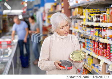 Elderly woman reading labels on tin cans with canned food in store 126201184