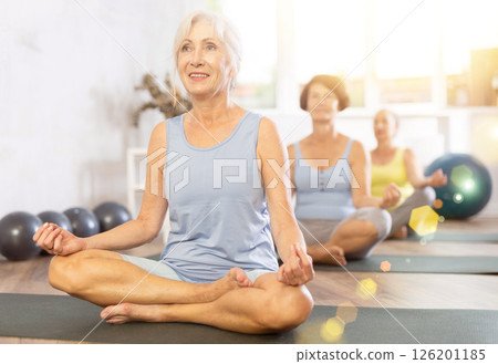 Elderly woman in company of hatha yoga lovers performs half-lotus pose, Ardha Padmasana Elderly woman in company of hatha yoga lovers performs half-lotus pose, Ardha Padmasana 126201185
