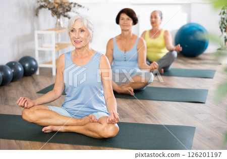 Elderly woman in company of hatha yoga lovers performs half-lotus pose, Ardha Padmasana 126201197