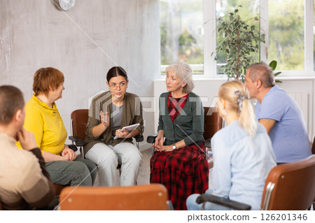 Young woman presides over support group meeting with mature people listening 126201346