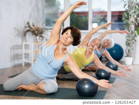 Old woman practicing half lotus pose with softball under arm in light fitness room 126201427