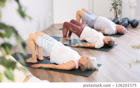 Elderly woman in company of hatha yoga lovers performs bridge pose, setu bandha sarvangasana Elderly woman in company of hatha yoga lovers performs bridge pose, setu bandha sarvangasana 126201575