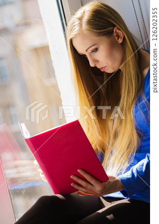 Woman sitting on window sill reading book at home Woman sitting on window sill reading book at home 126202355
