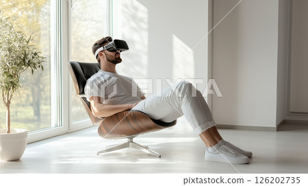 Man enjoying virtual reality while relaxing in a modern chair in a serene white room at home, experiencing peaceful moments alone 126202735