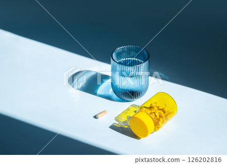 Glass of Water and Yellow Medicine Bottle with Pills and one Capsule Nearby in Hard Light. Taking Vitamin for Health and Wellness. Glass of Water and Yellow Medicine Bottle with Pills and one Capsule Nearby in Hard Light. Taking Vitamin for Health and Wellness. 126202816